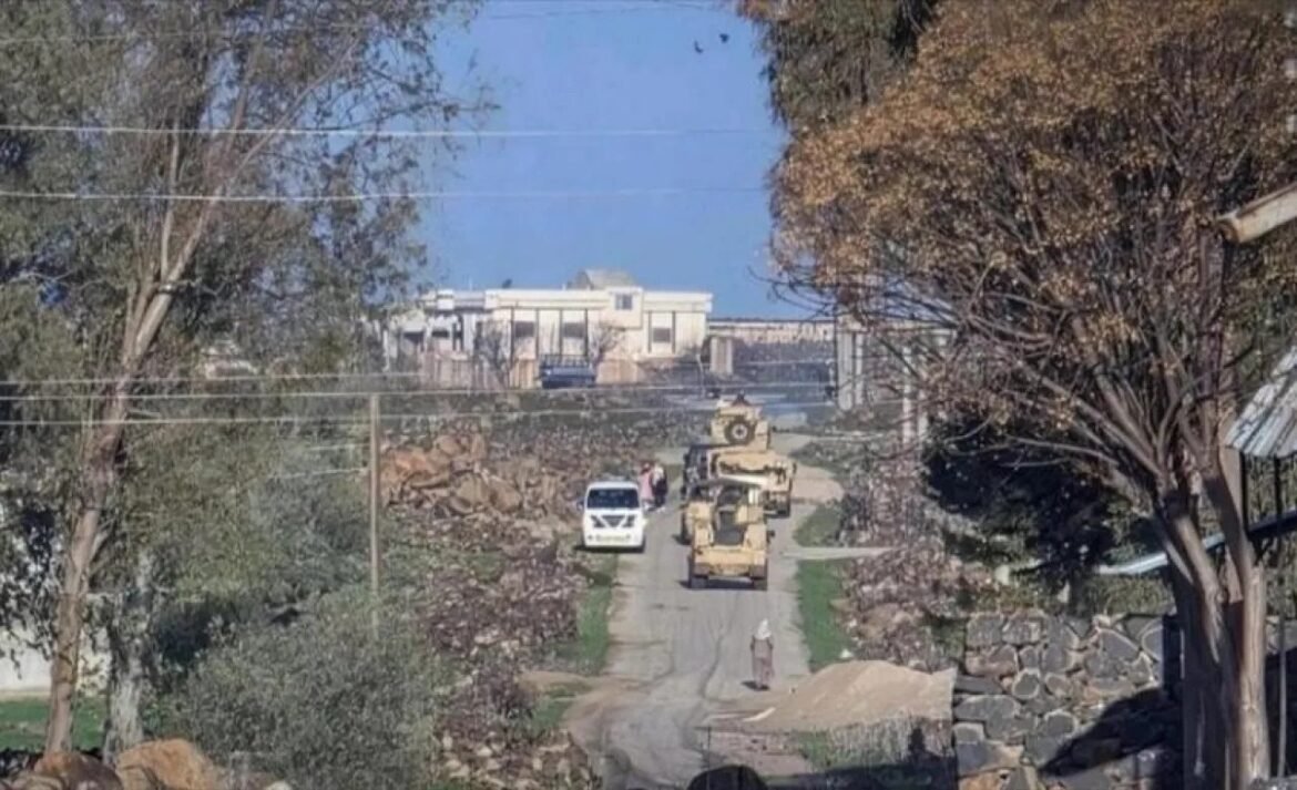 Line of beige armored vehicles driving single-file down a narrow residential road, with a white van nearby and trees along the sides.