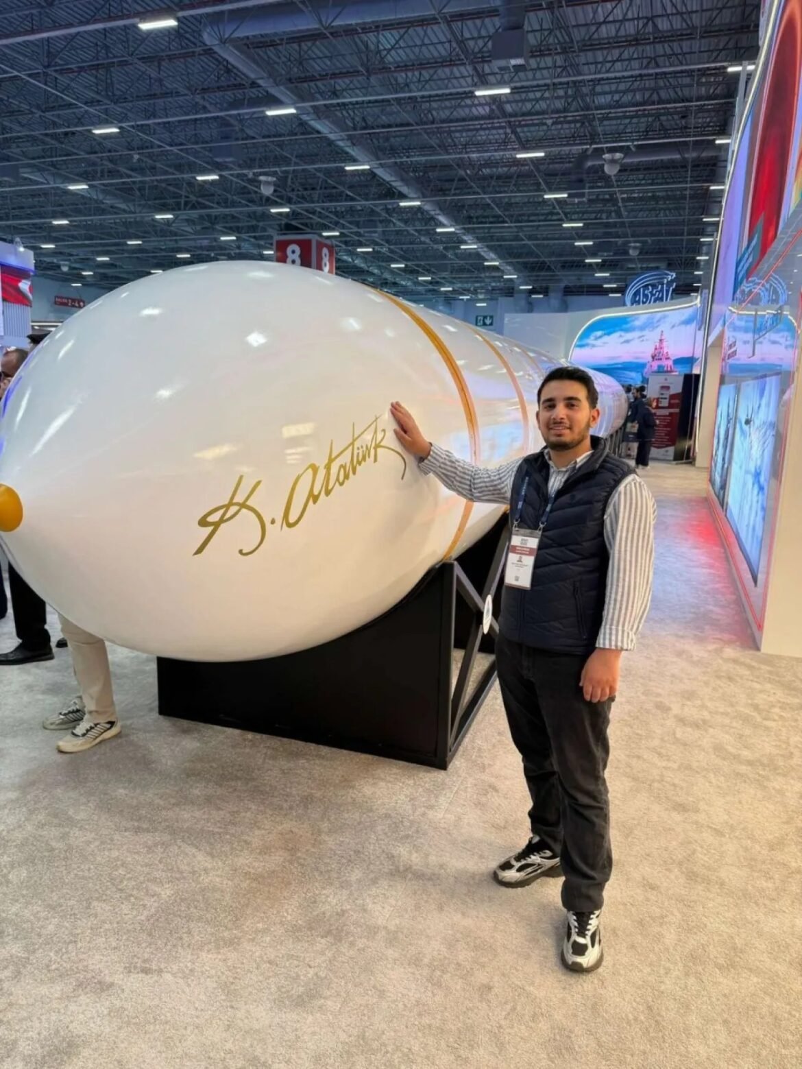 Man at a trade show touching a giant white capsule-shaped exhibit with a gold signature, in a busy hall.