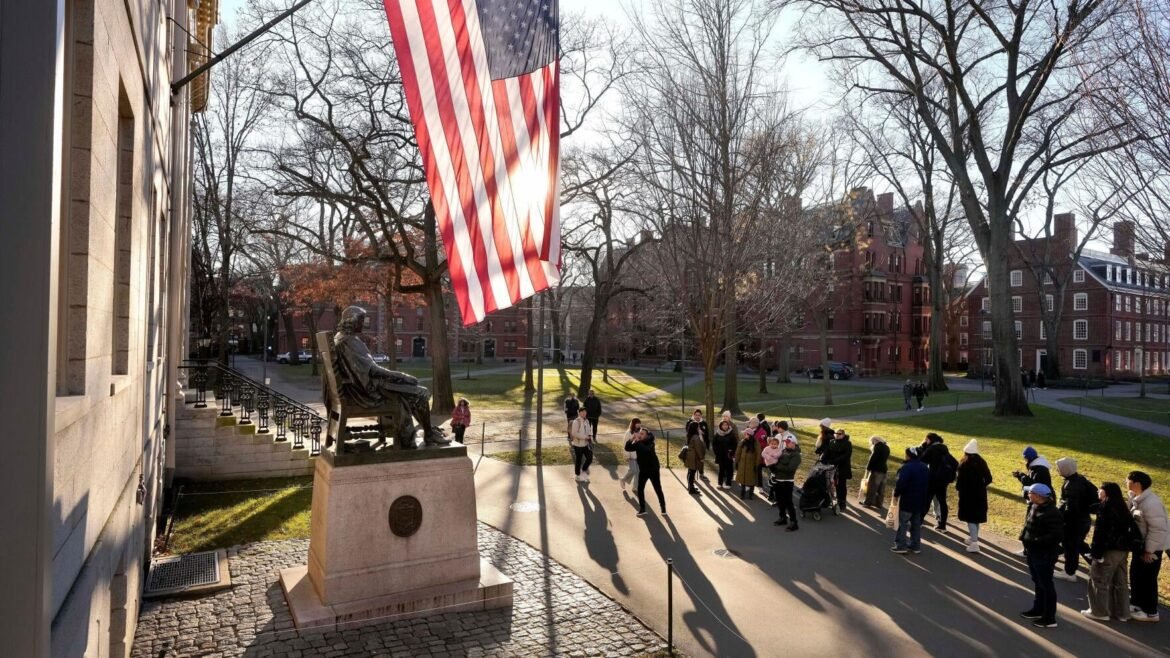 American flag hanging from a stone building beside a seated bronze statue on a pedestal, with a campus crowd and bare trees in the background.