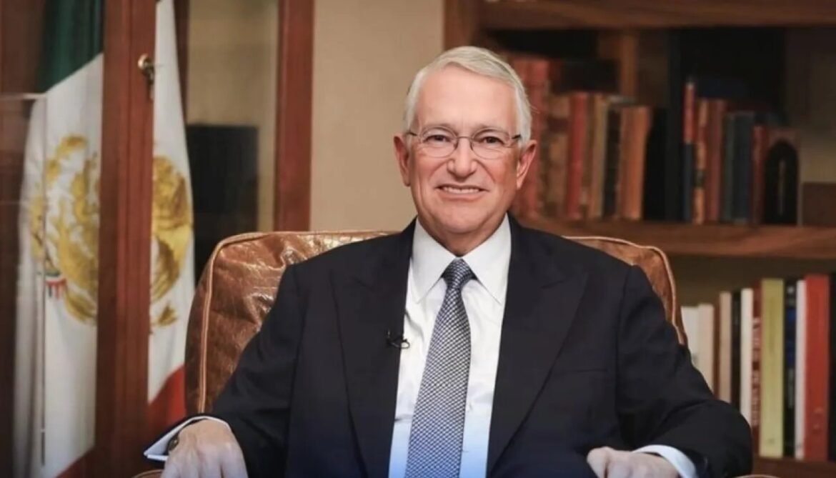 Smiling older man in a dark suit and tie seated in an office, with a bookshelf and Mexican flag behind him.