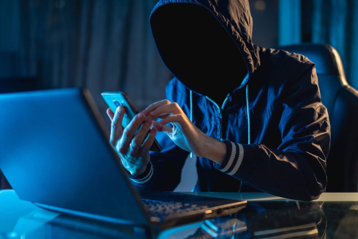 Person wearing a hoodie types on a smartphone while sitting at a glass desk with a laptop nearby, dim blue lighting suggestive of hacking activity