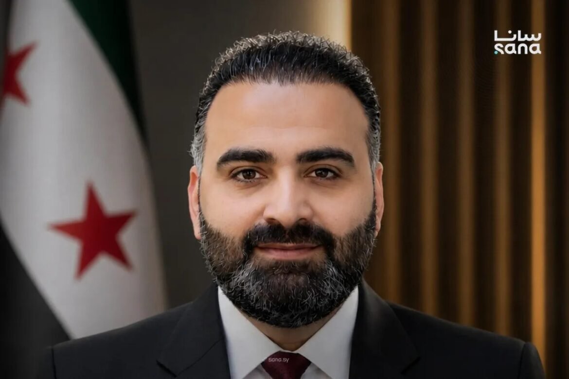Professional head-and-shoulders portrait of a man with a dark beard in a suit and burgundy tie, with a flag featuring red stars in the background and a Sana logo in the corner.