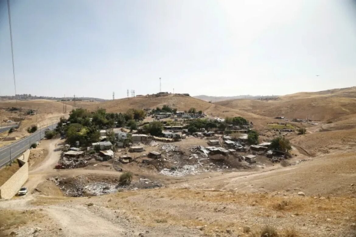 Small arid hillside village with scattered ruined stone huts and dusty roads, center shows a cluster of trees and debris around collapsed structures.