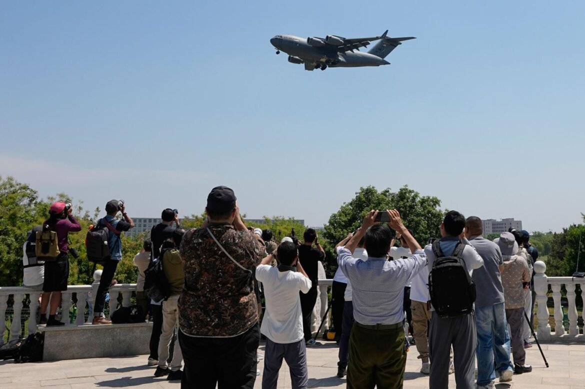Crowd of people on a viewing platform taking photos of a large plane flying overhead against a clear blue sky.