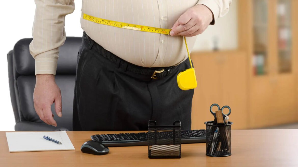Man in an office stands at a desk, measuring his waist with a yellow tape measure around his midsection.