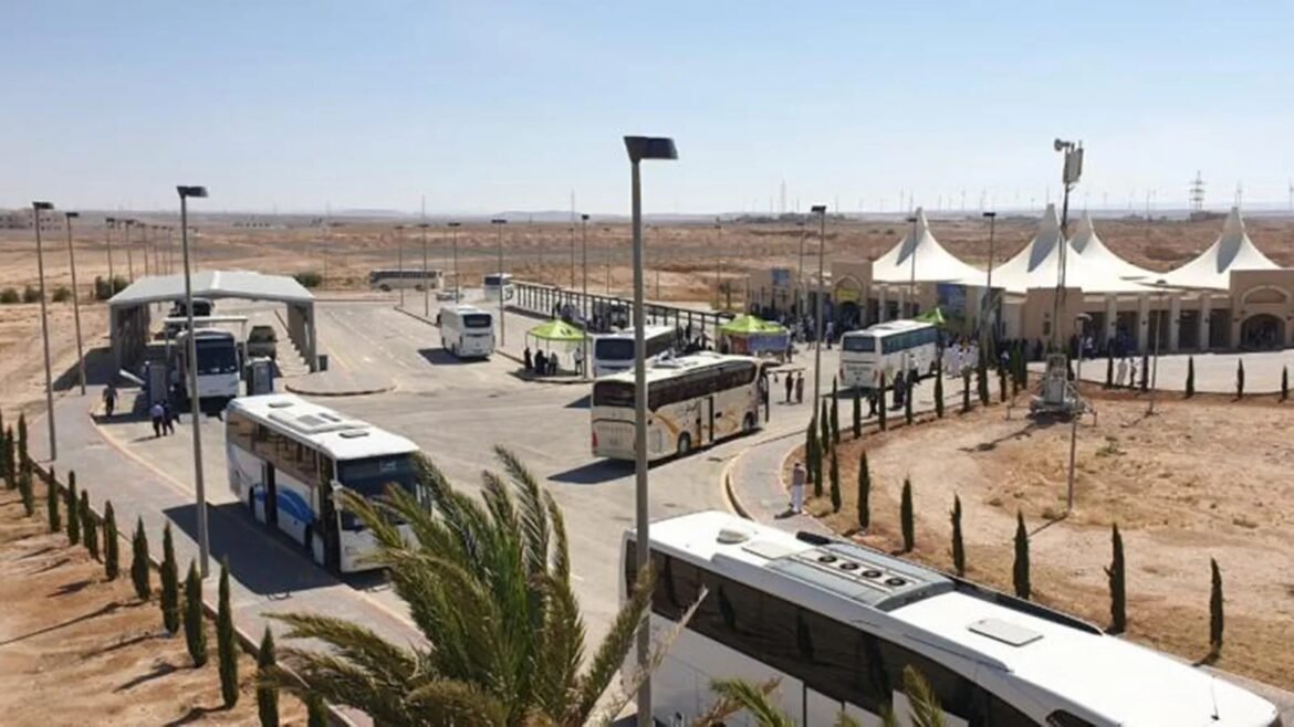 Bus depot in a desert landscape with several white coaches parked at bays and white tented buildings in the distance, people nearby.**