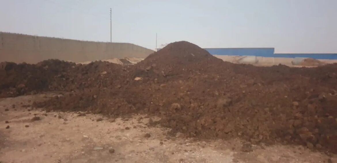 Large mound of brown soil in an outdoor construction yard with a long beige wall and a blue building in the background.