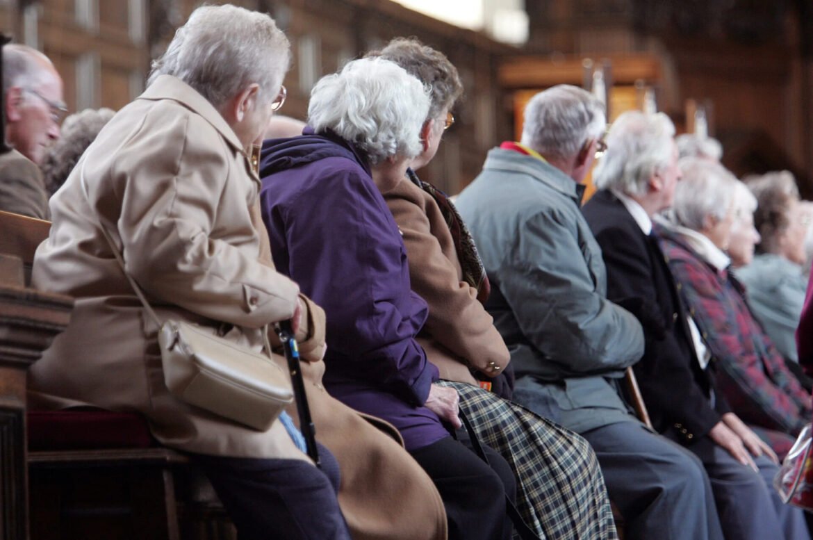 Senior adults seated in a church pew, wearing coats, facing to the right and watching a service.