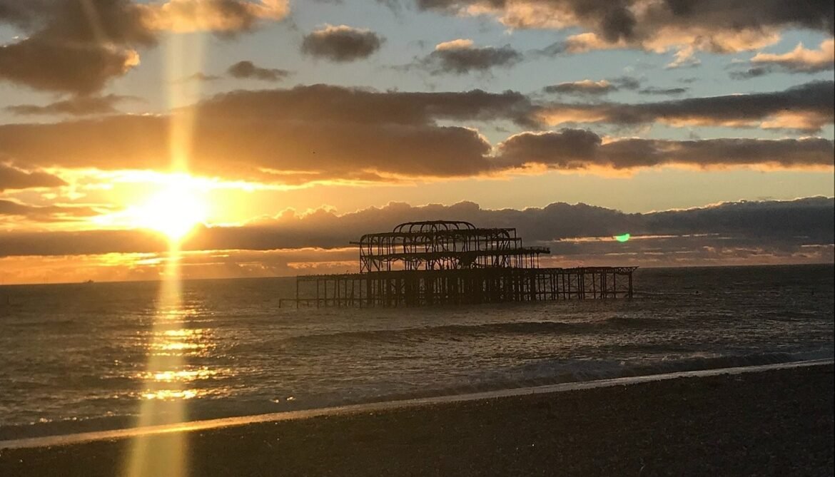 Sunset over the calm sea with a silhouetted rusted pier extending into the water; golden light reflecting off the waves.