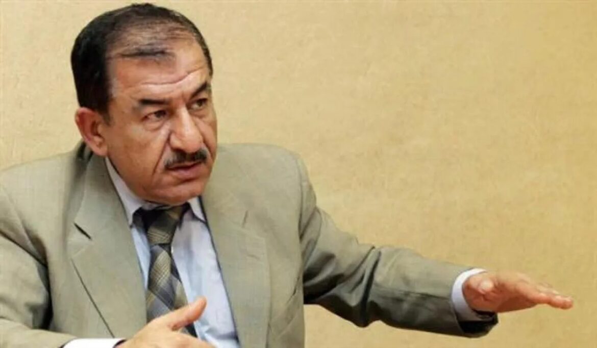 Older man in a light suit gestures with his right hand as if explaining something in a meeting setting.