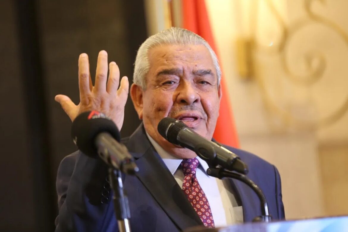 Older man in a suit and red tie speaks at a podium, raising his left hand as he addresses the audience. A microphone is in front of him and a flag is visible in the background.