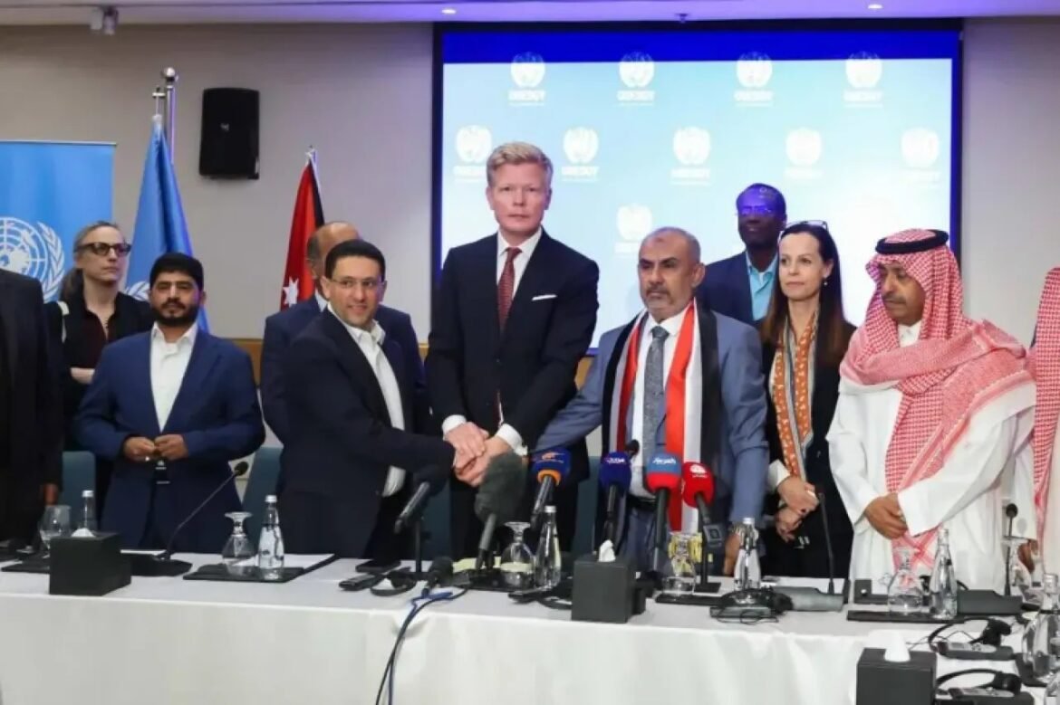 Group of officials in a conference room greet with a handshake over a table of microphones, UN backdrop behind them.