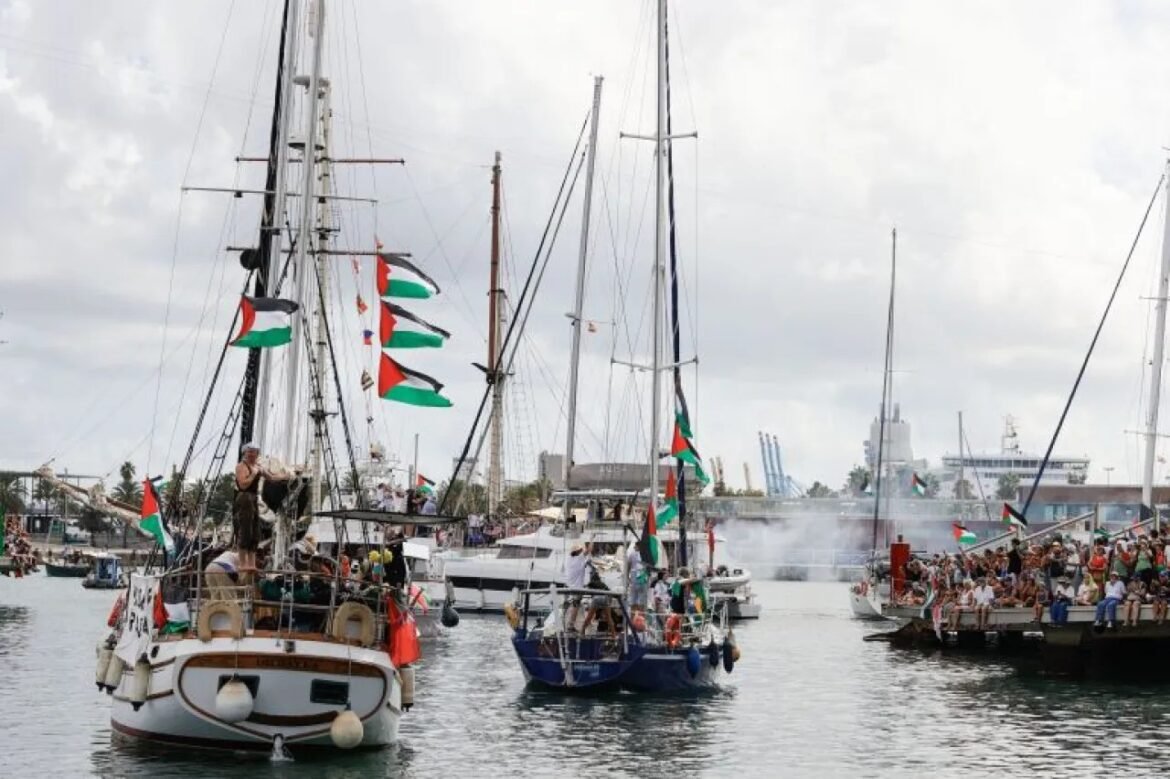 Sailboats with tall masts and green, white, and red flags parade through a harbor as crowds watch from the dock.
