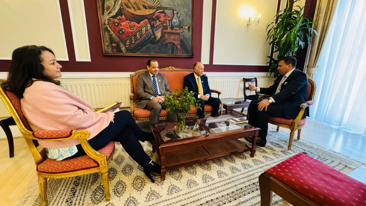 Four people in a formal living room seated for a meeting: a woman in a pink wrap on the left, two men on a red upholstered sofa, and a man in a dark suit across from them.