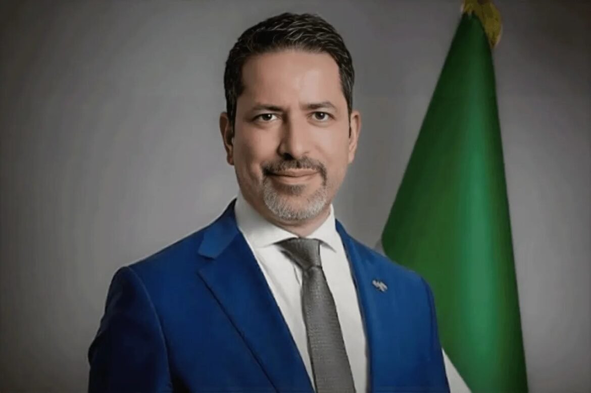 Portrait of a man in a blue suit and gray tie with a green flag in the background.