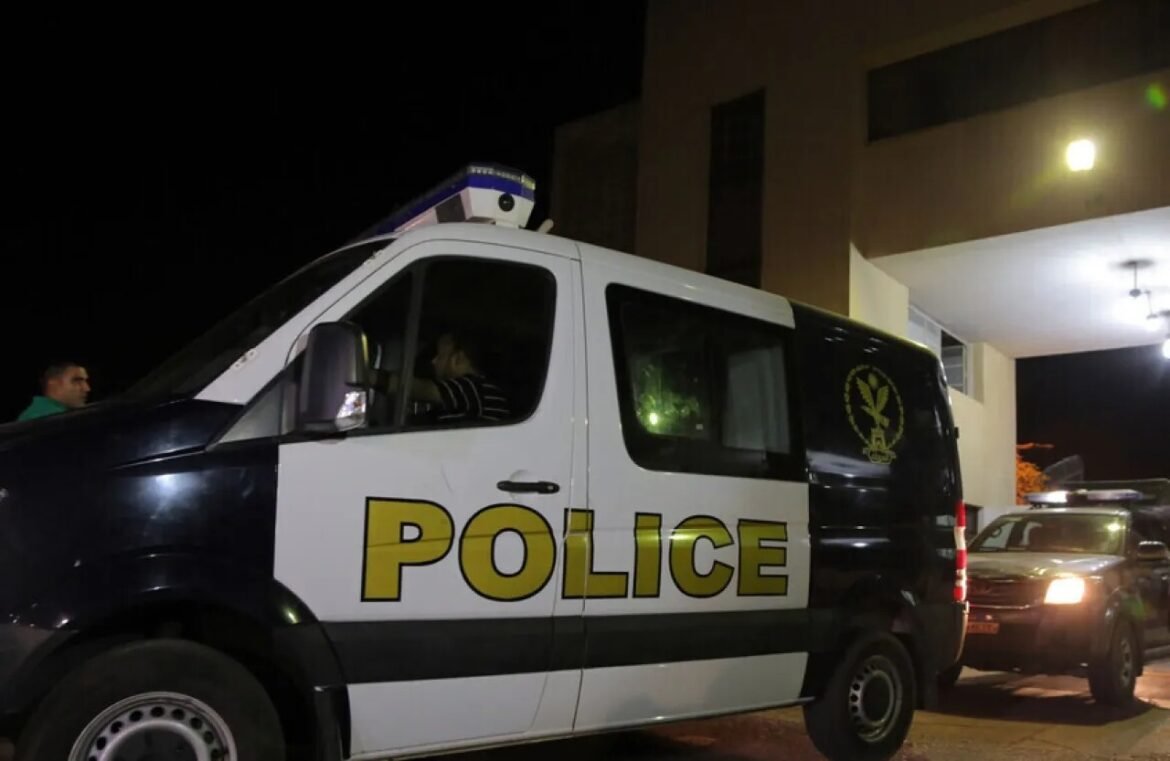 Night scene with a white police van parked beside a building; large yellow 'POLICE' lettering on the side and a crest on the rear window. A dark car is behind it.