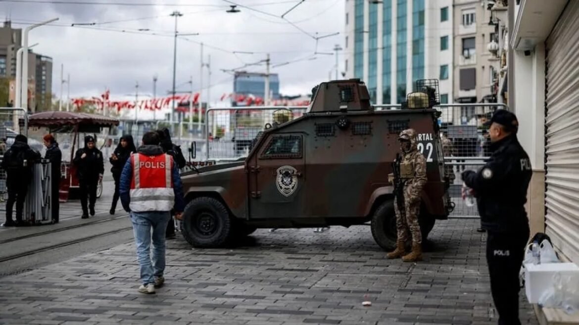 Armed armored vehicle and several police officers guarding a city street with barricades and pedestrians nearby.