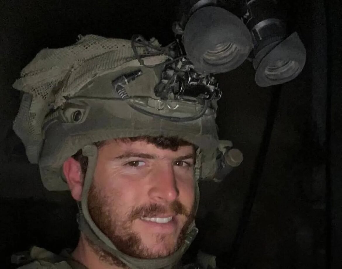 Male soldier wearing a helmet with mounted night-vision goggles, smiling at the camera in a dim setting.