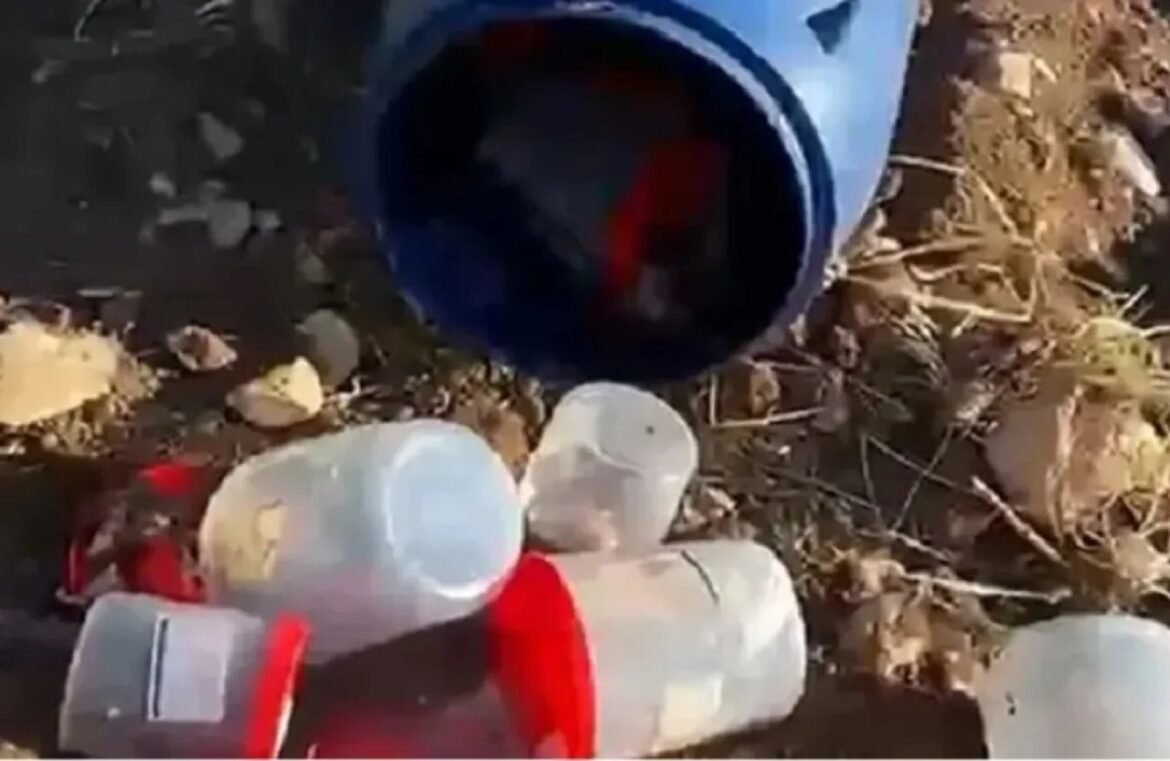 Blue plastic bucket on its side with several empty translucent bottles and red caps scattered on dirt ground, indicating littered area.