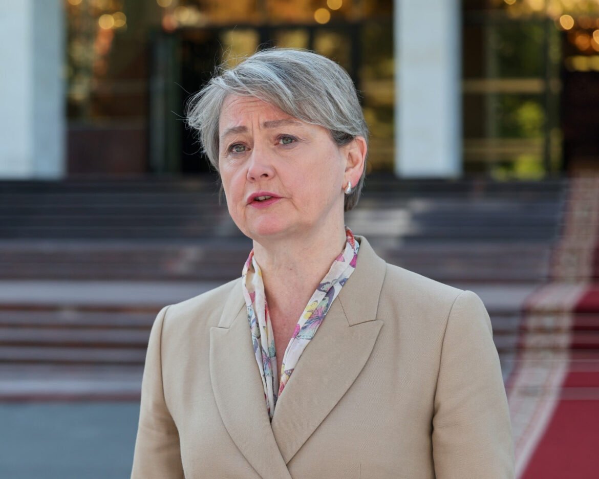 Portrait of a middle-aged woman with short gray hair, wearing a beige coat and floral scarf, outdoors near steps and a building.