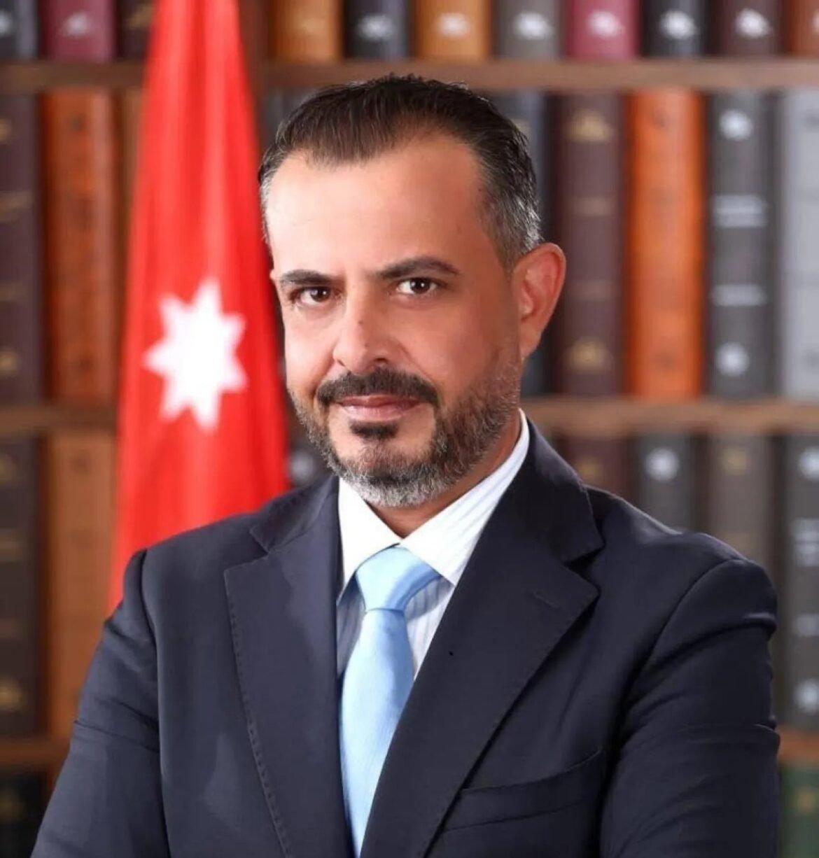 Professional portrait of a man in a dark suit and light blue tie, with a bookshelf and Turkish flag in the background.