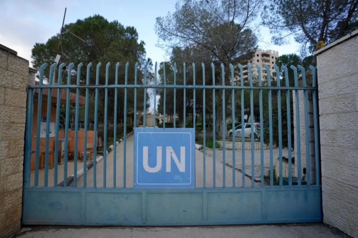 Blue UN gate with a large UN sign; security checkpoint and planted trees behind the barrier.