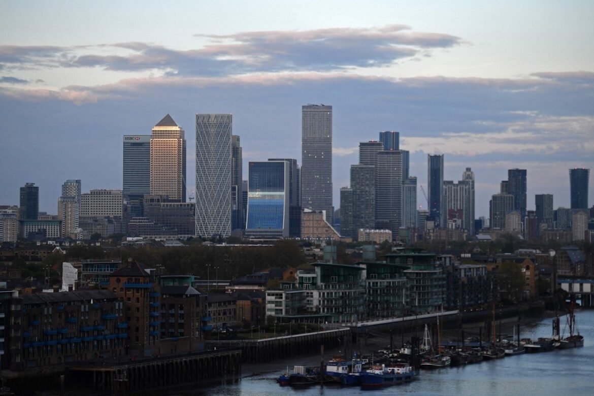Canary Wharf skyline in London viewed across a river, with modern high-rises and a marina in the foreground under a cloudy sky.