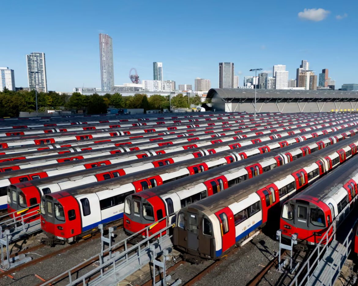 Row upon row of red-and-white metro trains parked in a rail yard with a city skyline in the distance under a blue sky.