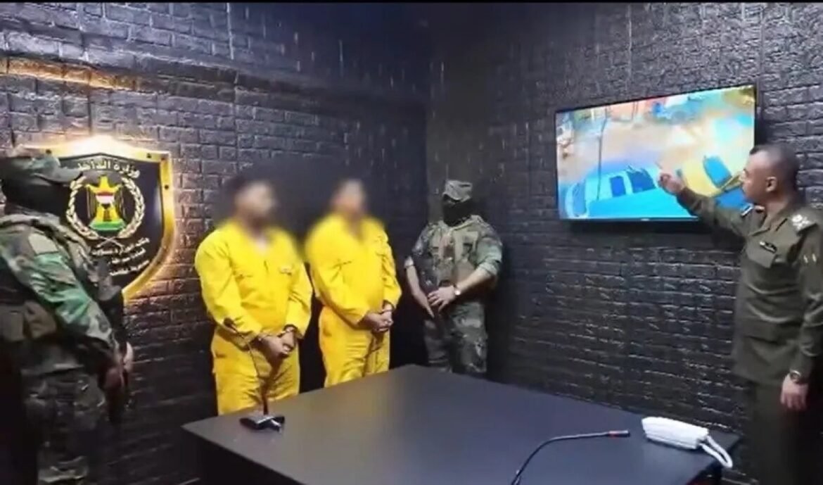 Two detainees in yellow jumpsuits with hands cuffed stand between armed guards in a dark brick-walled room as a uniformed officer points at a wall screen.
