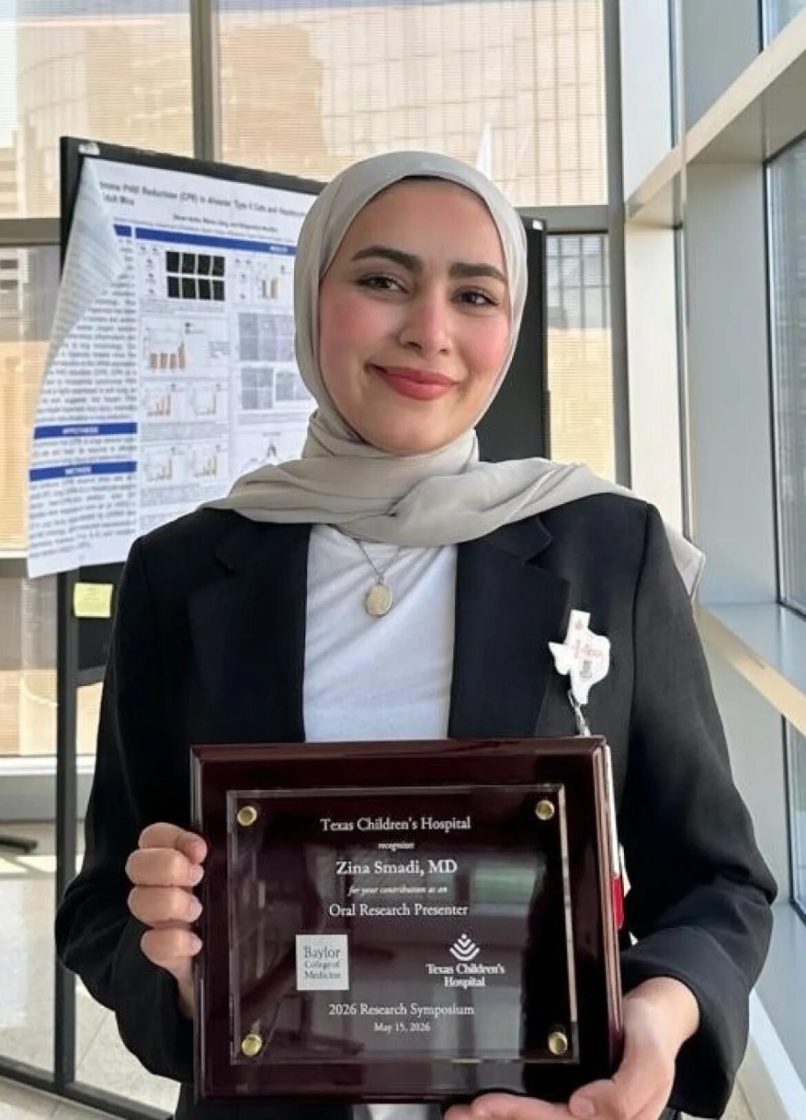 Smiling woman in a light hijab and blazer holding a glass award in front of a conference poster.