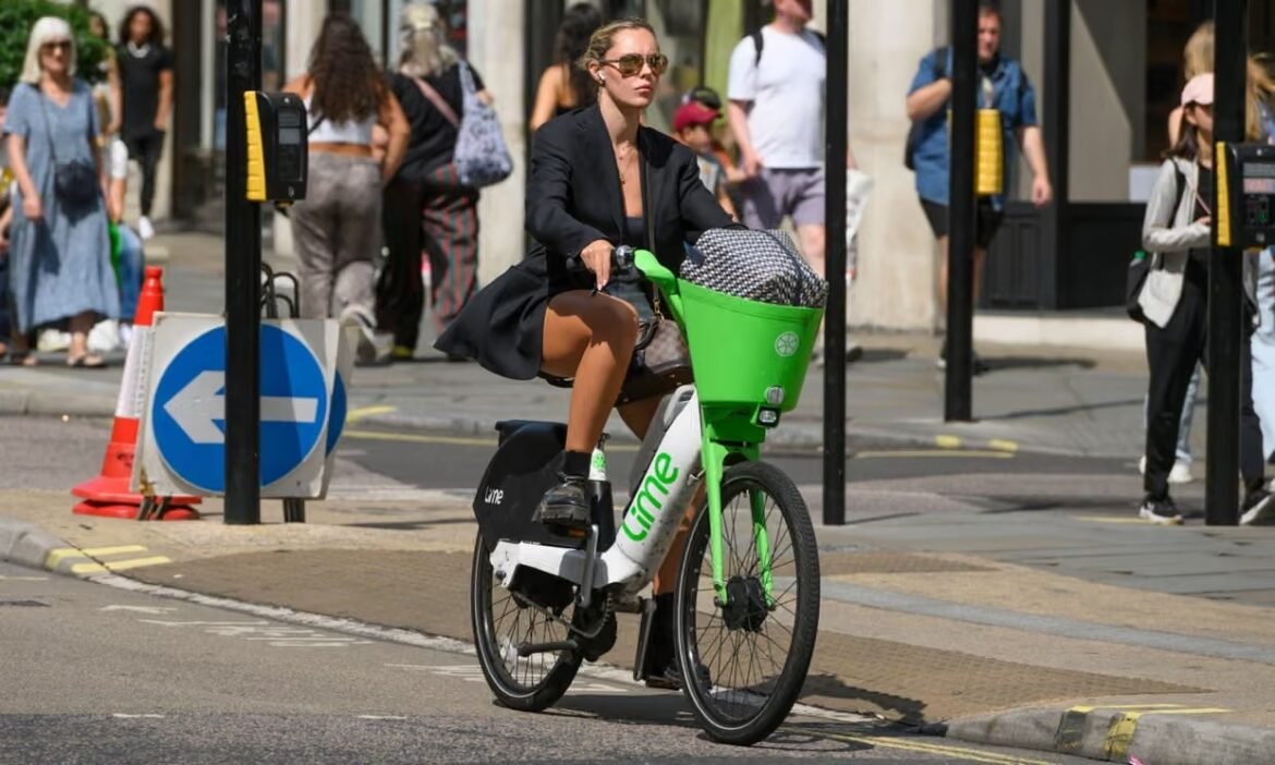Woman in black blazer riding a lime-green bike with a basket through a city street, pedestrians in the background.
