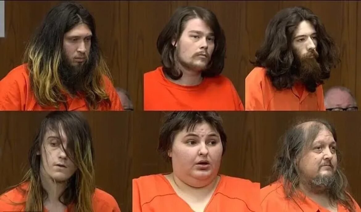 Six people in orange jail jumpsuits seated in a courtroom setting, facing forward in a lineup.