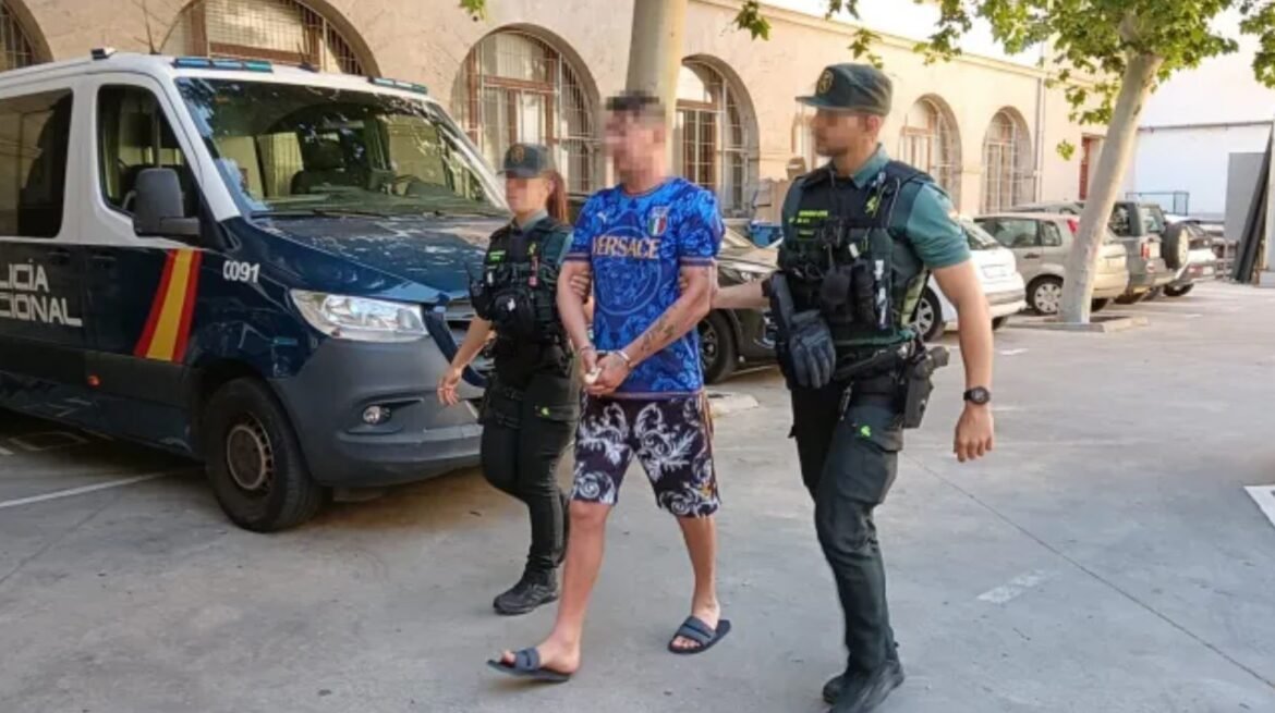 Two police officers escort a handcuffed man in blue shirt and patterned shorts past a police van in a street setting.