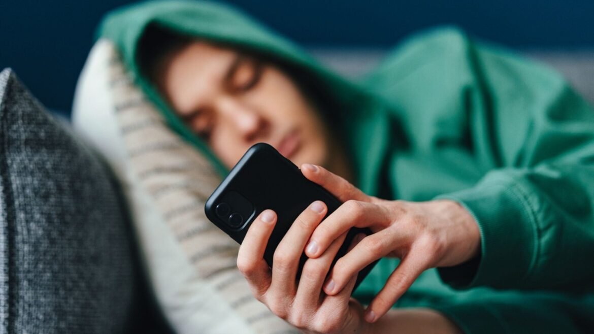 Person lying on a couch, wearing a green hoodie, holding a smartphone in both hands close to their chest.