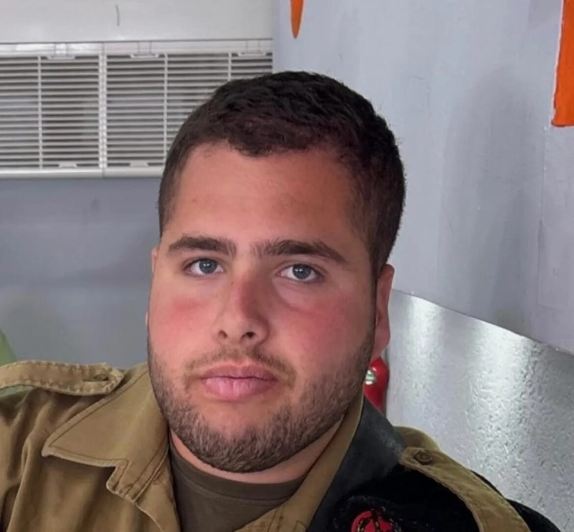 Close-up of a man with short dark hair and stubble wearing a tan uniform, looking at the camera.