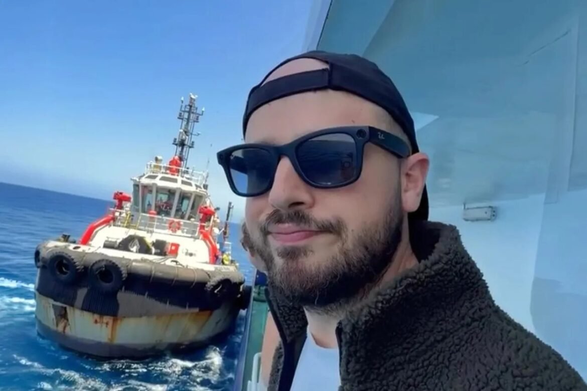 Man wearing a cap and sunglasses on a deck, with a tugboat pushing beside a blue sea and clear sky behind him