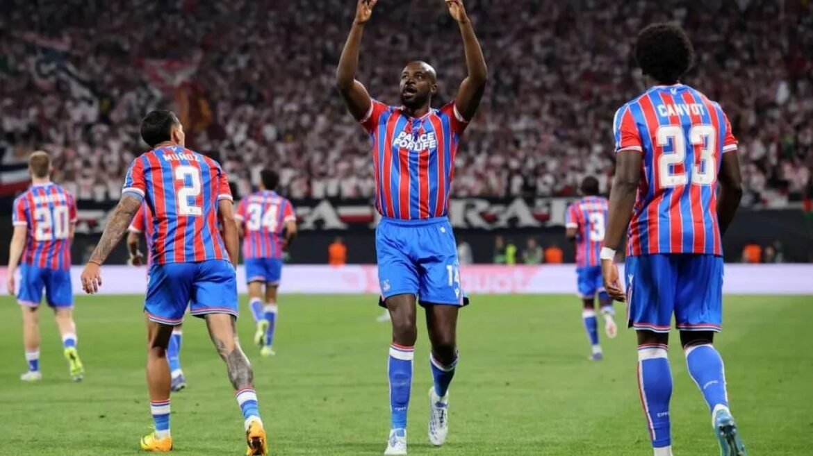 Soccer players in red-and-blue striped kits celebrate on the field as fans cheer in the stands.