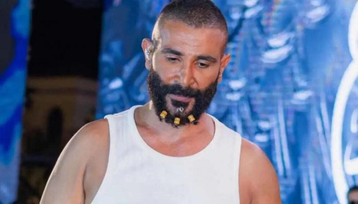 Bearded man in a white sleeveless shirt wearing bead accessories on his beard, looking down on a blue-lit stage backdrop.