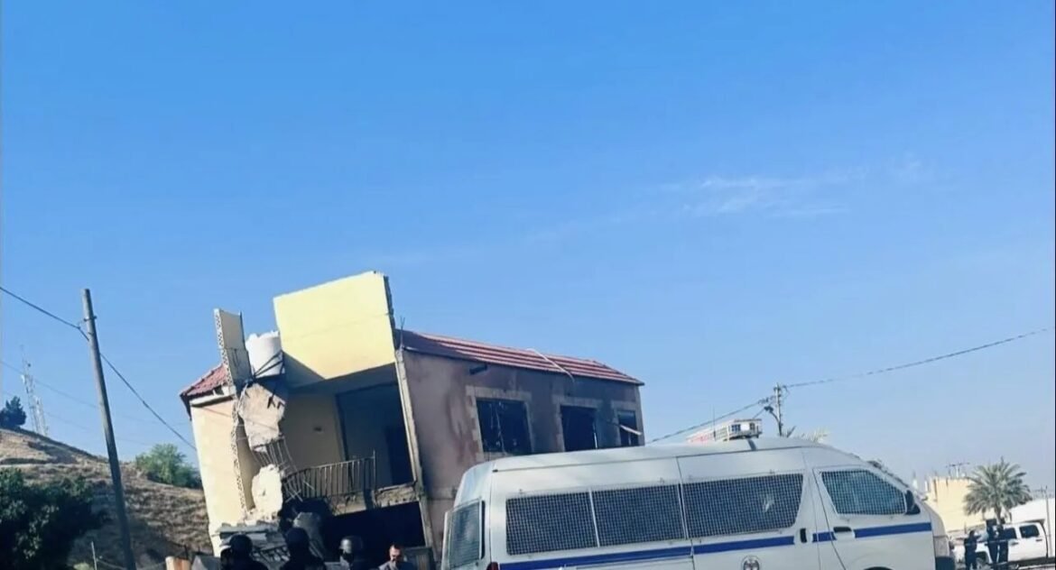 Partially collapsed two-story building with debris and a white van with blue stripes parked in front under a clear blue sky.
