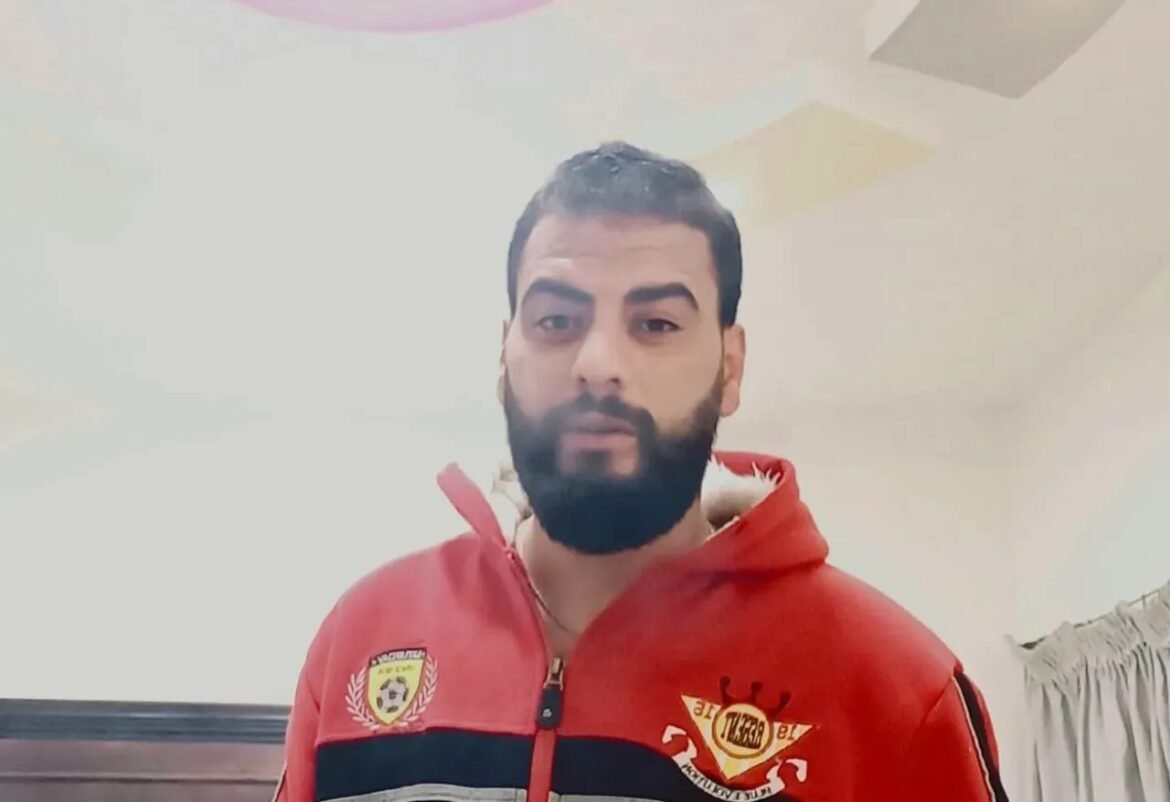 Man with dark hair and beard wearing a red zip-up hoodie with patches, indoors, looking at the camera.