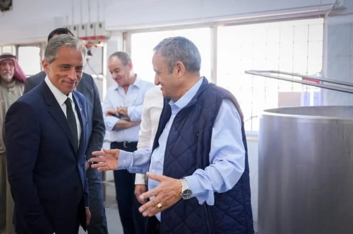 Two men in a factory talk near large stainless steel equipment, one gesturing with his hands while the other listens attentively, with several people in the background.