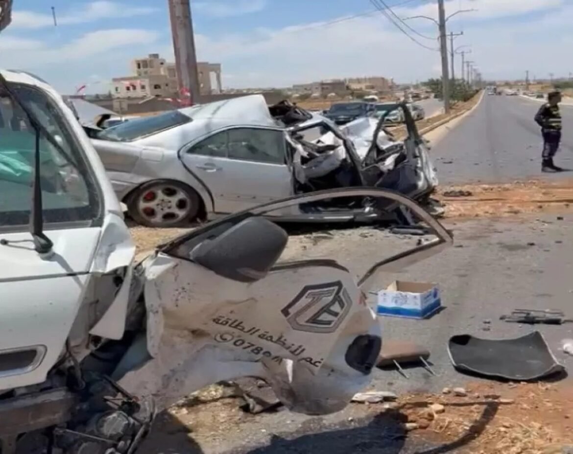 Rubble and crushed cars at a roadside crash, debris scattered across the road with a damaged vehicle in the foreground and a police officer in the distance.