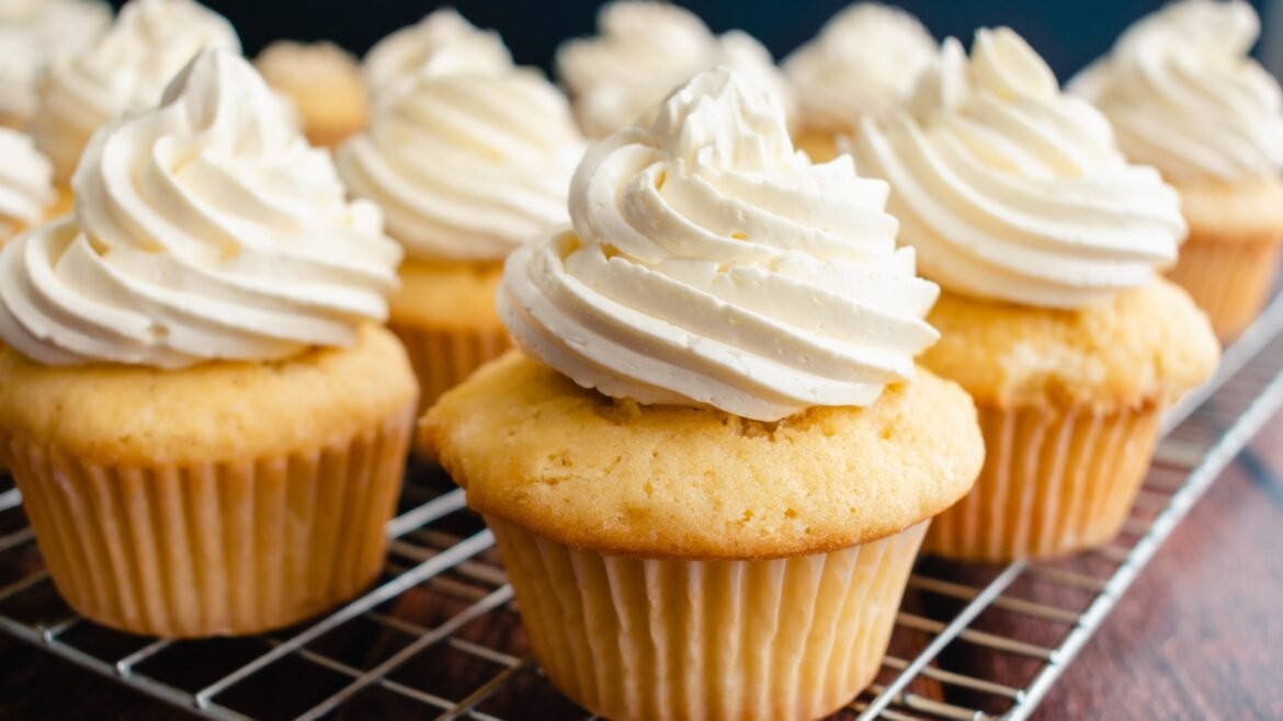 Cupcakes with swirled white frosting cooling on a wire rack in a bakery setting.