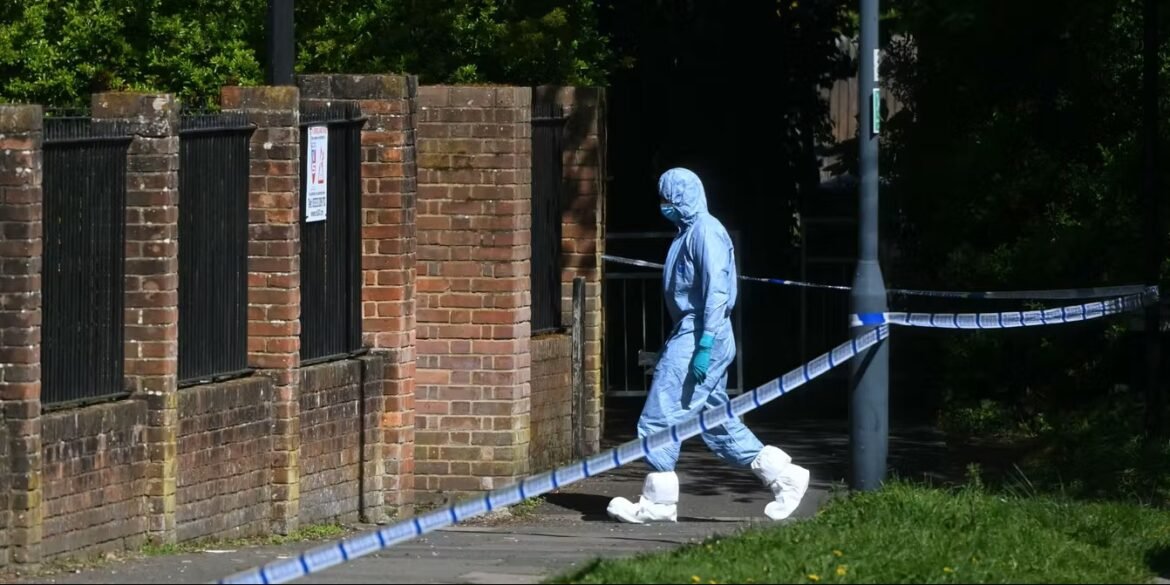 Person in a blue hazmat suit and gloves walking along a taped-off sidewalk beside a brick wall, suggesting a restricted area.