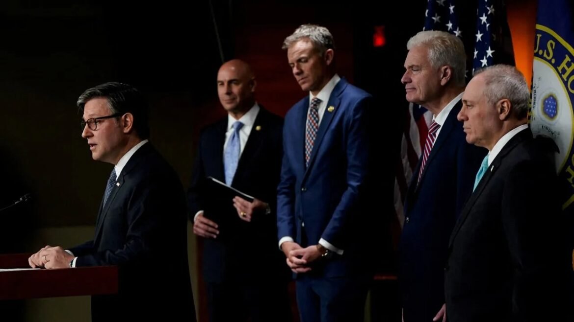 Man in a dark suit speaks at a podium as four suited men stand behind him during a press conference, with flags in the background.