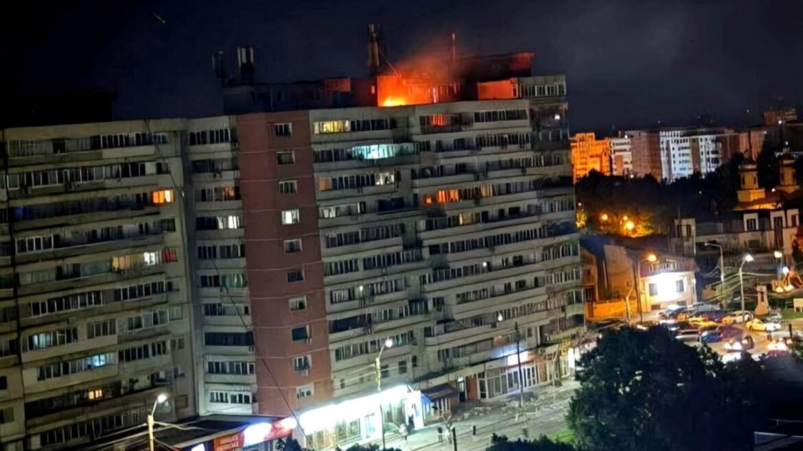 Nighttime view of a multi-story apartment building with visible flames and smoke on the roof on fire at the top floor area, city lights around it.