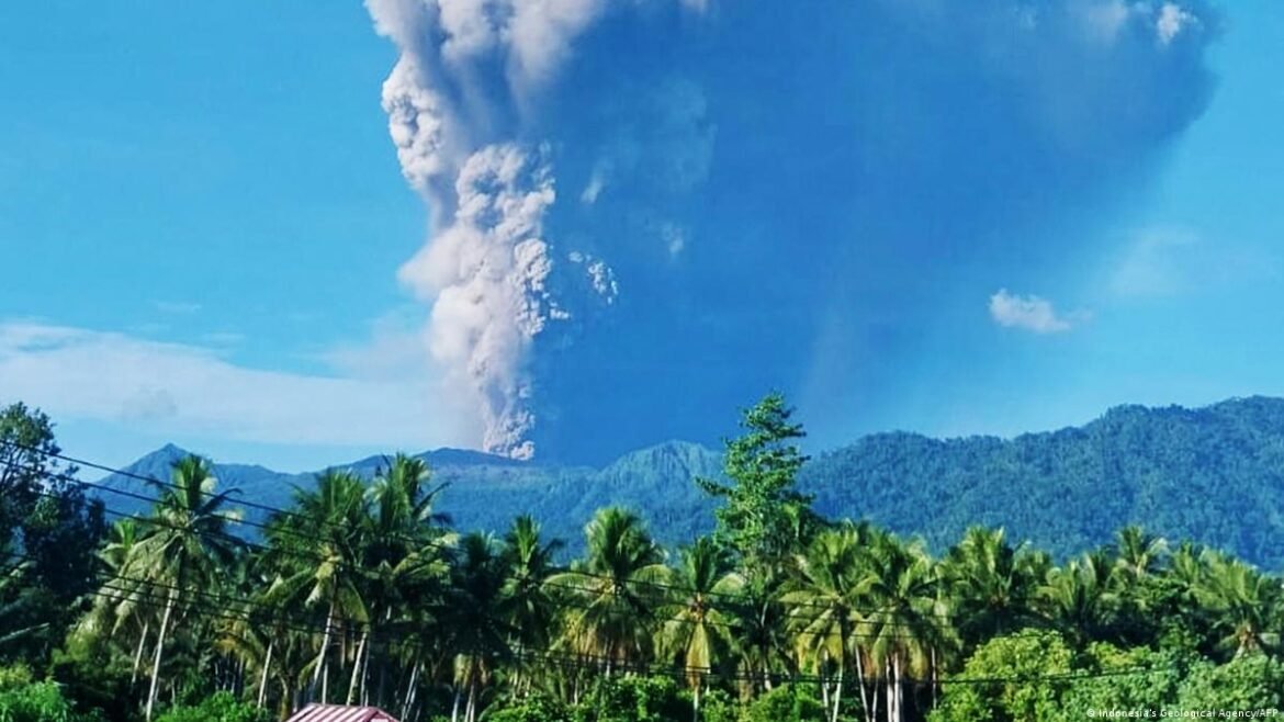 Volcano erupting with a tall ash plume rising over green hills and a row of palm trees in the foreground.
