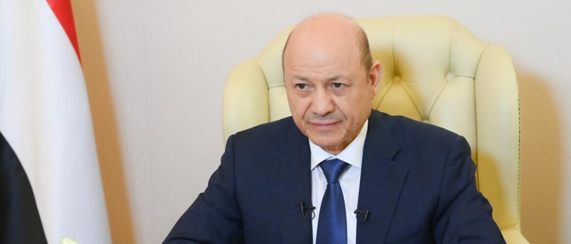 Bald man in a dark blue suit and blue tie sits at a desk in a cream leather chair, with a flag visible on the left.