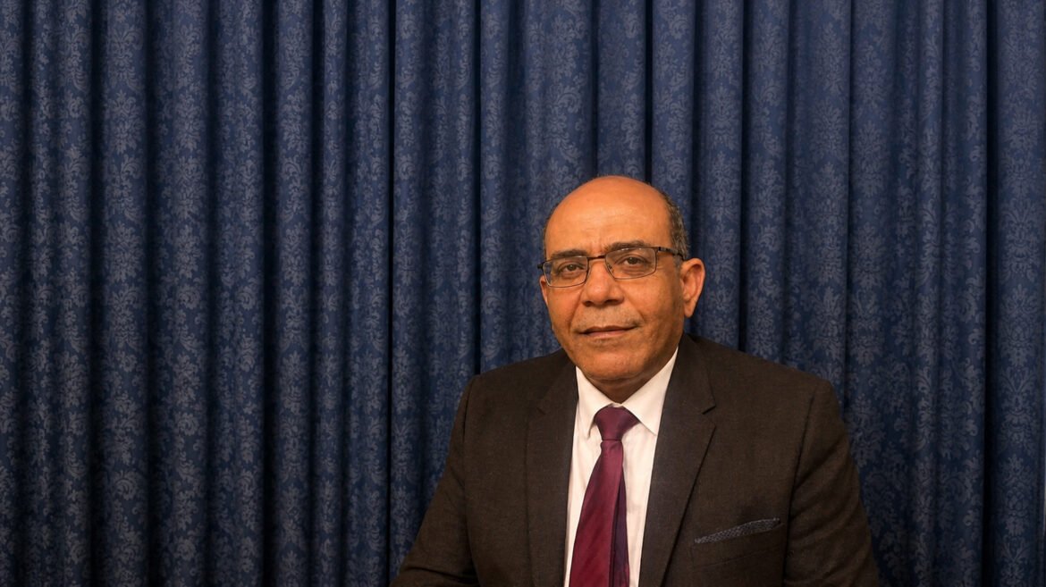 Smiling man in a dark suit and burgundy tie sits before a blue patterned curtain backdrop.