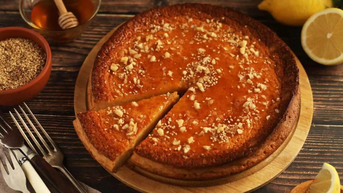 Round almond-topped cake on a wooden board with a slice cut out, lemons and honey nearby on a rustic table.